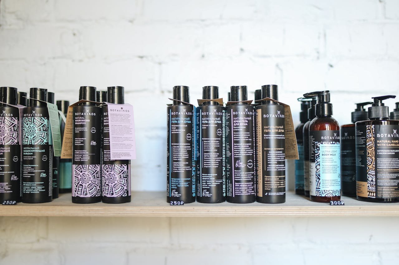A variety of natural cosmetic bottles lined up on a wooden shelf against a white brick wall, suggesting eco-friendly beauty.